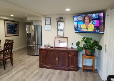 A living space with a mounted TV displaying a news anchor, stainless steel refrigerator, wooden sideboard with decor, dining table on left, and potted plant on the right.