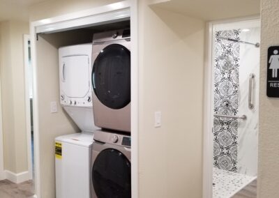 A laundry area with stacked washer and dryer units is adjacent to a bathroom with a white tiled floor and black floral pattern. A restroom sign is on the wall beside the bathroom entrance.