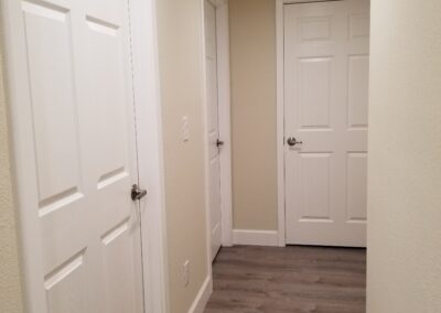 A hallway with three white doors and beige walls. The door at the end of the hallway is numbered 5. The floor is covered with wood-like vinyl planks.