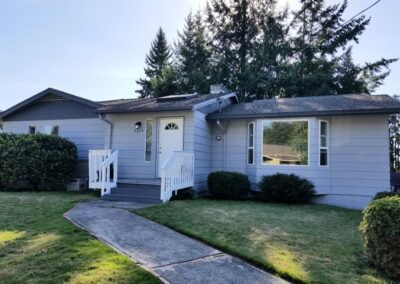 A small, single-story house with light gray siding, white trim, and a white front door. The house has a front porch with white railings, a concrete pathway, and a lawn with bushes and trees.