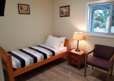 A small bedroom with a wooden single bed, white and patterned bedding, a nightstand with a lamp, a purple chair, and two framed pictures on the wall. A window with blinds is on the right side.