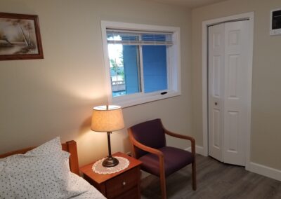 A small bedroom with a bed, a nightstand with a lamp, a purple chair, a window, and a closet with white folding doors. A framed picture hangs on the wall above the bed.