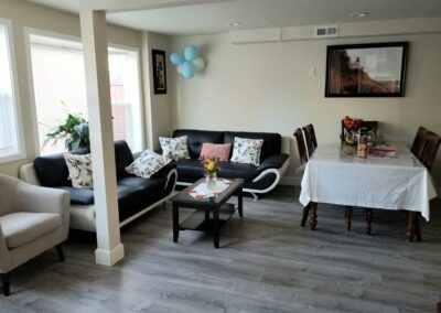 A living room with modern furniture, including a black sofa with patterned cushions, a coffee table, a beige armchair, and a dining table with chairs and a white tablecloth. A few wall decorations are visible.