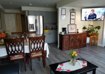 A kitchen and dining area with a wooden dining table, chairs, and cabinets. A TV is mounted on the wall displaying a man. There are houseplants, and a vase with flowers on a coffee table.