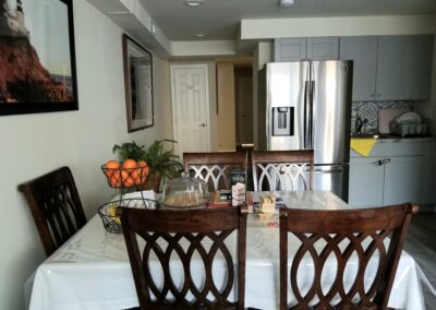 A kitchen area with a dining table set for four. The table has a white cloth, a fruit basket with oranges, a cake under a cloche, and a few cardboard boxes. Stainless steel refrigerator in the background.