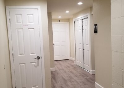 A hallway with light-colored walls and wood flooring has three white doors. One door on the right has a restroom sign. Ceiling lights illuminate the area.