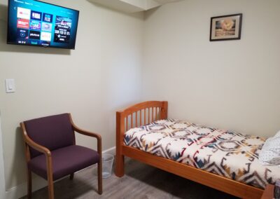 A small, tidy room with a wooden single bed, a blanket with a geometric pattern, a purple armchair, a wall-mounted TV displaying streaming service icons, and a framed picture on the wall.