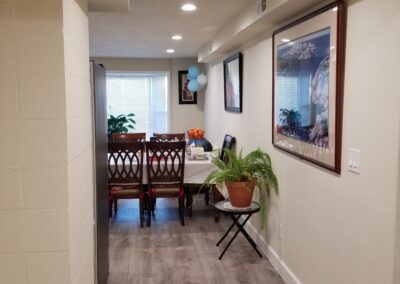 A narrow hallway leads to a dining area with a table and chairs. Walls are decorated with framed pictures, and a potted plant sits on a small stand to the right.