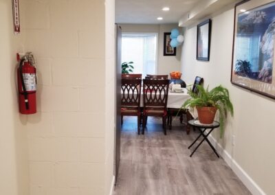 A hallway with light wood flooring leads to a dining area with a table, chairs, a potted plant, and blue balloons. A fire extinguisher is mounted on the left wall. Framed pictures hang on the right wall.