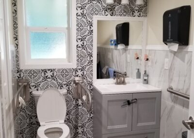 A bathroom featuring a toilet with grab bars, a sink with a mirror, patterned wallpaper, and a frosted window. The floor is made of wood-like material.