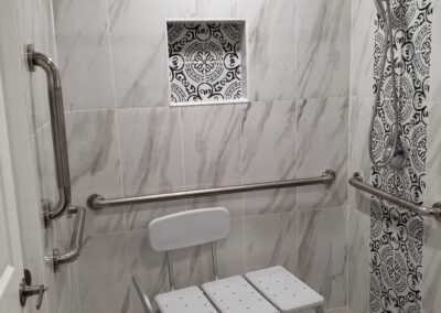 A shower equipped with a white chair, grab bars, a handheld showerhead, and a decorative tiled wall.