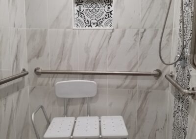 A tiled shower with a bench seat, grab bars, and a handheld showerhead. The shower area has a patterned tile niche and pebbled flooring.