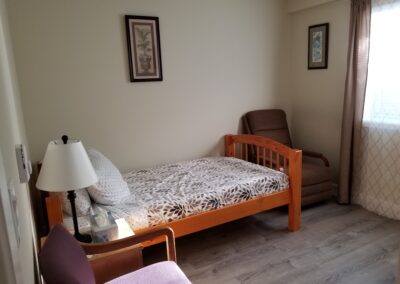 A simple bedroom with a single bed, pillow, chair, recliner, side table with a lamp, and framed pictures on the wall. The room has a window with closed curtains allowing some natural light.