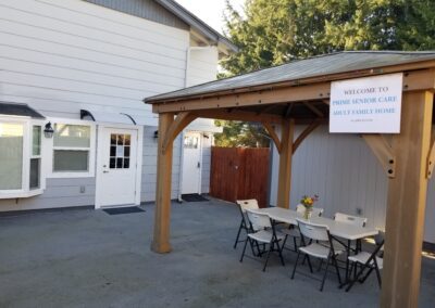 Covered outdoor seating area with folding tables and chairs, beside a house with a sign that reads "Welcome to Prime Senior Care Adult Family Home.