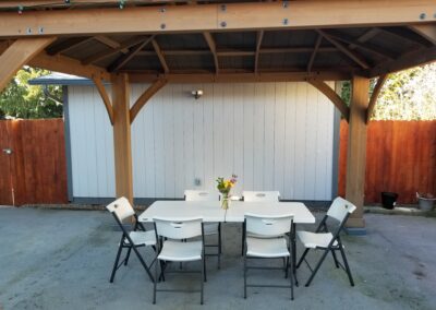 An outdoor patio under a wooden gazebo featuring a white rectangular table with a bouquet of flowers, surrounded by six foldable chairs. A wooden fence and white shed are in the background.