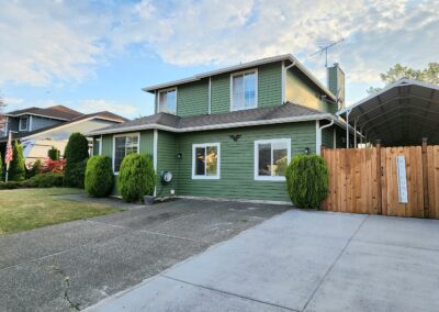 A green two-story house with white trim, a concrete driveway, wooden fence, and a covered carport. Lush bushes and a lawn are in the front yard under a blue sky with some clouds.