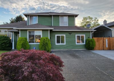 A green two-story house with a brown roof, white trim, and a paved driveway. The front yard features a red shrub, green bushes, and a wooden fence on the right.