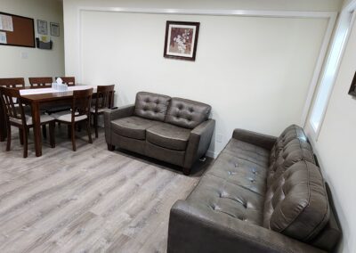 A living room with a dining table and six chairs, two leather couches, framed artwork, and a bulletin board on the wall, all set on a wooden floor.