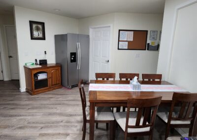 A modern kitchen with hardwood flooring features a dining table with six chairs, a stainless steel refrigerator, a small wooden sideboard, and wall-mounted frames and notice boards.