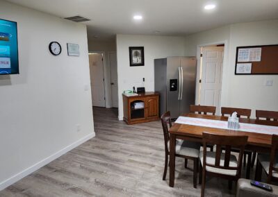 A living area featuring a wall-mounted TV, a clock, a dining table with six chairs, and a refrigerator in the background. The table has a decorative runner, and two water bottles.