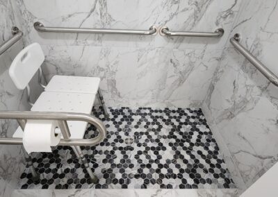 A marble-tiled shower with safety grab bars, a white shower chair, a handheld showerhead, and a mounted toilet paper holder. The floor has black and white hexagonal tiles.