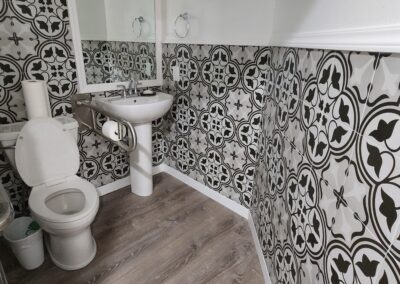 A small bathroom featuring patterned black and white tiled walls, a toilet with a grab bar, a pedestal sink, a wall mirror, and a white paper towel dispenser. The floor is wooden.