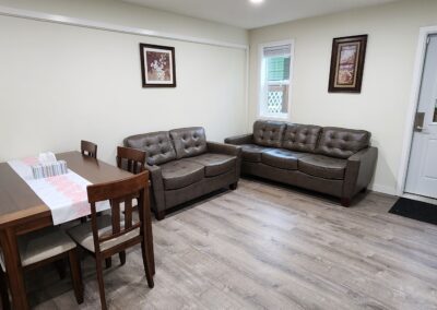 A living room with a dining area, featuring a brown leather sofa set, a wooden dining table with chairs, and wall art. The room has light-colored walls, wood flooring, and a door to an outside area.