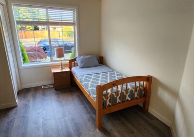 A small bedroom with a single bed next to a nightstand located under a window. The room has hardwood flooring, light-colored walls, and a framed picture on the wall. A car is visible outside the window.