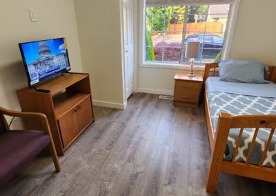 A small, tidy bedroom features a single bed, a chair, a TV on a wooden stand, and a window overlooking a fenced yard.