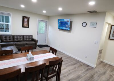 A living and dining area with a rectangular wooden table, six chairs, a brown sectional sofa, wall-mounted TV, framed pictures, and a wall clock. Walls and ceiling are light, with recessed lighting.