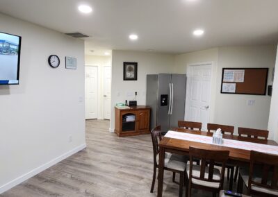 A small dining room with a wooden table and six chairs, a TV mounted on the wall displaying a news broadcast, a refrigerator, a sideboard, and a clock. The room has light-colored walls and hardwood flooring.