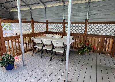 A covered outdoor patio with a white table surrounded by six white folding chairs. The area is enclosed by lattice panels and wooden railing, with potted flowers placed near the corner.