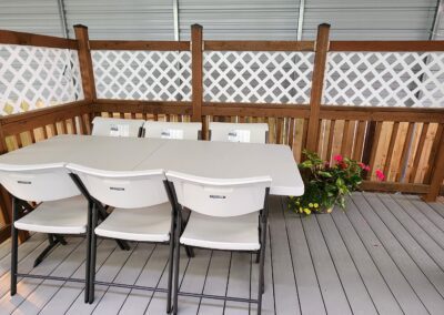 A small covered deck area with a white plastic table and five white folding chairs. A potted plant with pink flowers sits in one corner. The space is enclosed with wooden lattice fencing.