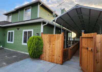 A green two-story house with a covered carport and a wooden fence. The property has neatly trimmed bushes and a cement driveway. The sky is partly cloudy.