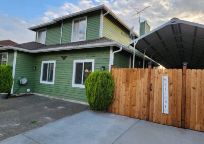 A green two-story house with white trim, a wooden fence, and a metal carport. The fence has a "WELCOME!" sign. Two large bushes are near the windows, and concrete paths are in front.